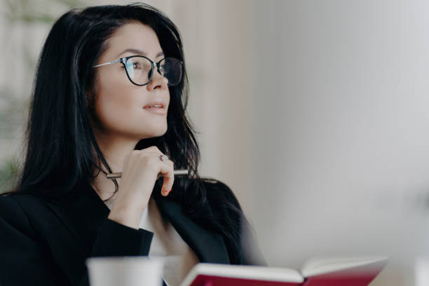 istock Thoughtful woman notes ideas, plans, and goals in organizer with glasses on, deep in thought. 1560333556