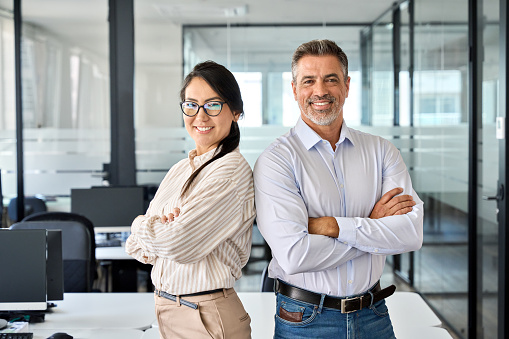 Happy confident professional mature Latin business man and Asian business woman corporate leaders managers standing in office, two diverse colleagues executives team posing arms crossed, portrait.