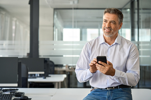 Happy mid aged business man using phone in office. Smiling mature confident businessman executive manager holding smartphone working on cell phone sitting at office desk looking away and thinking.