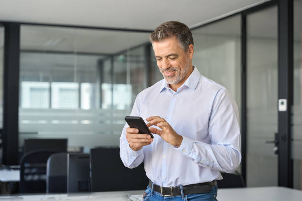 istock Smiling older business man executive standing in office using mobile phone. 1560356943