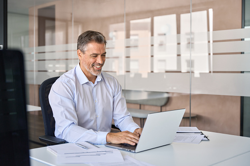 Smiling elegant handsome mid aged business man executive sitting at desk using laptop. Happy stylish professional businessman worker or manager working on computer technology in modern office.