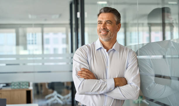 Happy mature latin business man standing in office arms crossed looking away. Happy mid aged older professional business man, mature latin executive manager, 50 years old investor standing in office arms crossed leaning at glass wall looking away thinking of success concept. piece of mind stock pictures, royalty-free photos & images