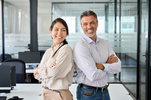 Happy smiling confident professional mature Latin business man and Asian business woman colleagues corporate managers standing in office, two diverse executives team laughing arms crossed, portrait.