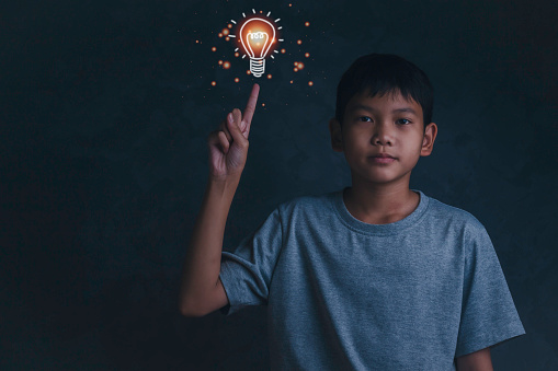 Boy is holding up his finger and a light bulb icon above it. Concept of child found solution, education, creativity and brainstorm isolated on gray wall background.