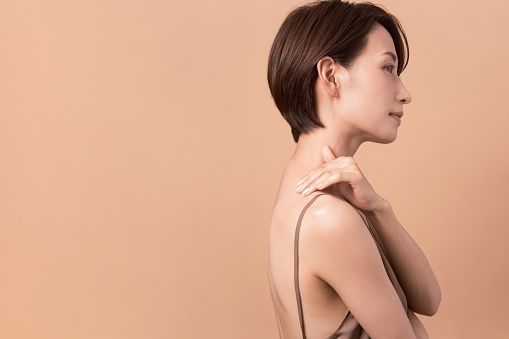 A beautiful Asian woman puts her hand on her shoulder. Sideways.  Studio shot, beige background.