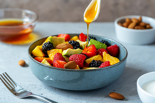 Healthy fruit salad in a grey bowl made with avocado, strawberry, raspberry, orange, blackberry, almonds and peaches. Teaspoon with honey dropping to the salad. Yogurt, honey behind. Grey background