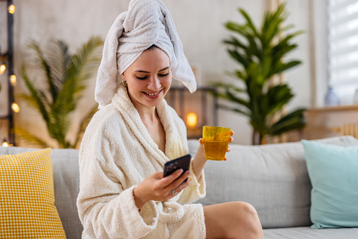 Beautiful young woman in a bathrobe sitting on the sofa in the living room, using her smart phone and drinking water.