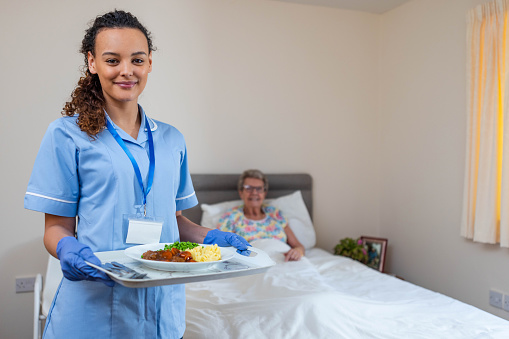 Three quarter length portrait of a community nurse visiting a senior woman who is lying in her bed at her home in Northumberland, England. The nurse is wearing her scrubs, smiling and looking at the camera while holding patient a hot cooked dinner on a tray.