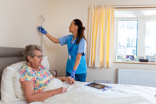 Medium view of a community nurse visiting a senior woman who is lying in her bed at her home in Northumberland, England.  The nurse is wearing her scrubs and attaching an IV drip to her patients hand.