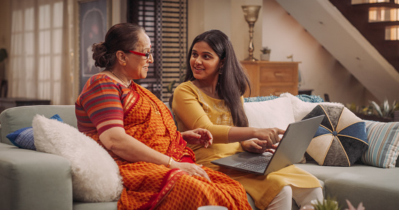 Beautiful Indian Mother and Daughter Use Laptop at Home: Browsing Websites for Clothing, Products Sales, Choosing Best for Online Shopping.Smiling Happy Ladies Enjoy Spending Family Time together