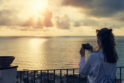 Teenage girl sightseeing Calabrian town of Tropea. She is filming sunset from the Belvedere Piazza del Cannone viewpoint. \nShot with Canon R5