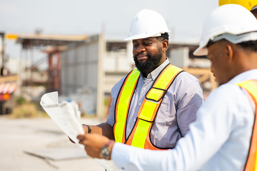 Happy Engineering worker in safety hardhat and architect looking at blueprint at industrial Heavy Manufacturing Factory. Prefabricated concrete walls. Professional Engineer Team working