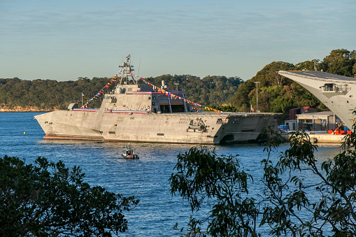 USS Canberra (LCS 30) and behind her, HMAS Canberra (L02), docked at Garden Island in Sydney Harbour, viewed through native vegetation.  USS Canberra is an Independence Class combat ship of the US Navy.  She is in port for a commissioning ceremony on 22 July 2023.  She is adorned with red, white and blue bunting and maritime flags.  HMAS Canberra is an amphibious assault ship of the Royal Australian Navy.  One person in camouflage uniform guards the stern deck.  An Australian Federal Police motorboat patrols the harbour.  Yellow buoys on the water mark the exclusion zone around the naval base.  This image was taken from Mrs Macquarie's Chair on a cold, sunny afternoon about 30 minutes before sunset on 20 July 2023.