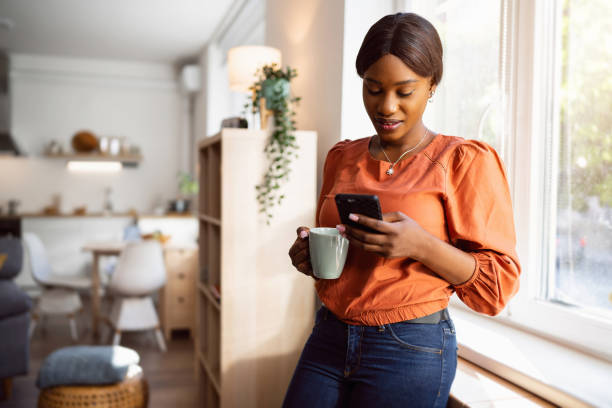 Young woman of Black ethnicity, using mobile phone while holding coffee