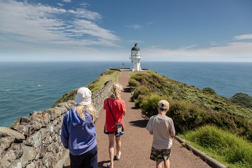 A mum and two children are walking away from the camera towards Cape Reinga lighthouse, which is at the far north of the North Island in New Zealand. The entire area is very important for the Maori people and they attach much spiritual and cultural significance to the area.