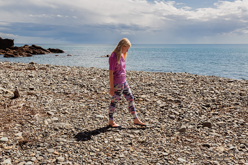 A young girl is walking on her own on a stony beach in deep contemplation. The sea is in the background with white clouds in the sky.
