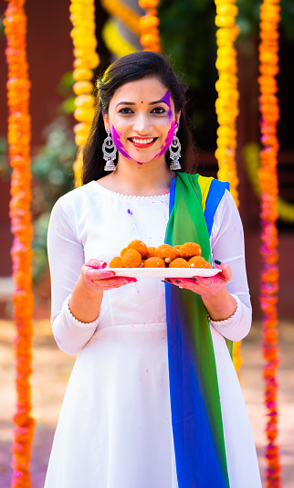Vertical shot of indian happy young girl with color face holding sweet or laddu by looking at camera on flower decorated background - concept of holi festival, happiness and joyful