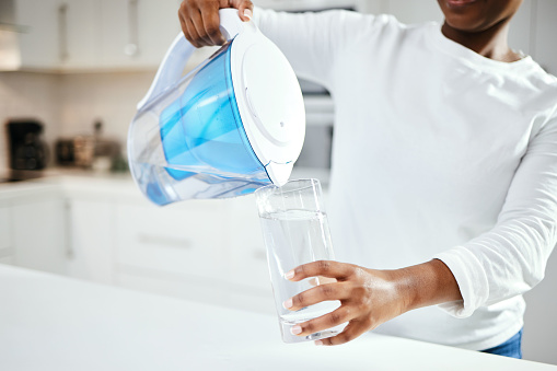 Person, jug and glass of filter water in kitchen for fresh drink, liquid and hydration. Closeup, thirsty woman or pouring pure aqua beverage in container for nutrition, drinking or filtration at home