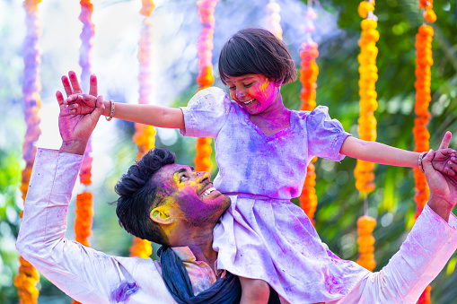 Indian father with his daughter on shoulder dancing during holi festival celebration on decoration flower background - concept of fatherhood, family bonding and enjoyment.