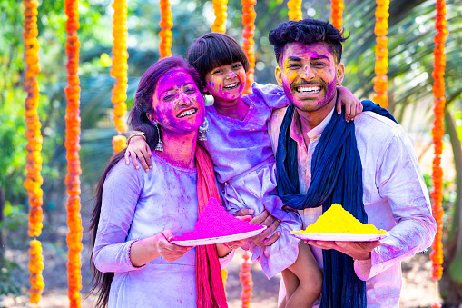 Young colorful indian couple with kid saying happy holi by looking camera - concept of festival greeting, wishes and family bonding.