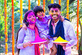 Young colorful indian couple with kid saying happy holi by looking camera - concept of festival greeting, wishes and family bonding