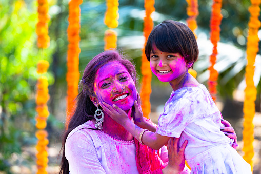 Happy young kid applying holi color on mother face by looking at camera during holi festival celebration