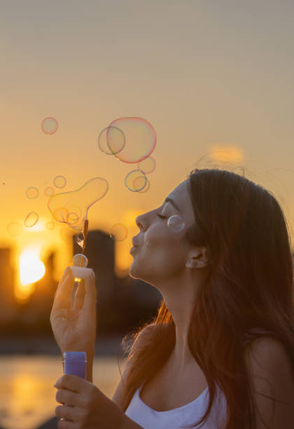 Pretty young woman is blowing bubbles front of cityscape at sunset.