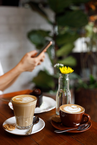A close-up of two cups of coffee and a sweet chocolate dessert cake on a cafe table with a woman using a smartphone in the background, waiting for her friend.