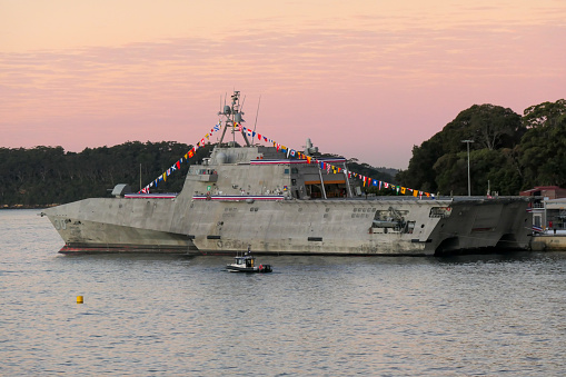 The Stars and Stripes are lowered in a ceremony on the USS Canberra (LCS 30), docked at Garden Island in Sydney Harbour. One person lowers the flag while two salute.  USS Canberra is an Independence Class combat ship of the US Navy.  She is in port for a commissioning ceremony on 22 July 2023.  She is adorned with red, white and blue bunting and maritime flags.  An Australian Federal Police motorboat patrols the harbour.  Yellow buoys mark the exclusion zone around the naval base.  This image was taken from Mrs Macquarie's Chair on a cold, sunny afternoon at sunset on 20 July 2023.