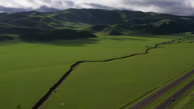 Aerial view of a central California valley with after a wet winter broke the drought. A highway with vehicles are seen. Green pastureland and meadows in a slow dolly drone shot with green hills, cloudy sky in background.