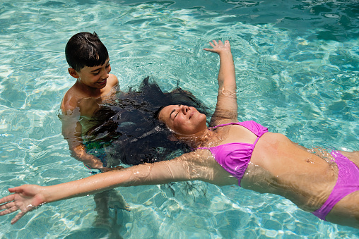 Mother and son in backyard pool in tropical setting. Kid is 10 years old and is wearing a blue swimsuit. Mom is in her fifties and wearing a pink bikini. Mother is floating on her back. Horizontal waist up outdoors shot with copy space. This was taken in Florida, USA.