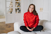 Relaxed woman in red sweater sits on sofa, holds phone, with New Year tree in living room.
