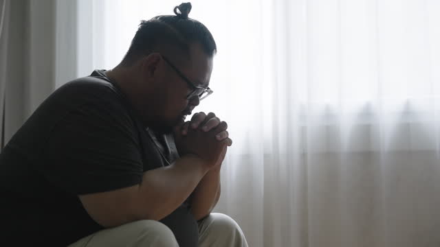 SLO MO: Side view of a chubby Asian guy sitting uncomfortably on the sofa in his living room with his head down and interlocking hands praying for the miracle to happen to him.