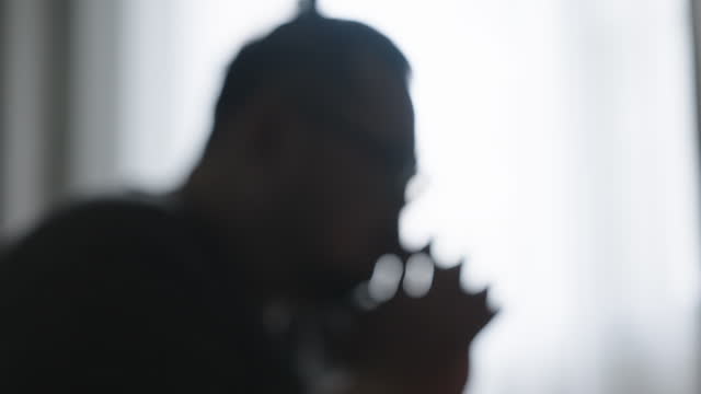 SLO MO: Blurred motion with bokeh effect and gradually becoming focus, close-up, side view of a moody plus-size Asian guy sitting on the sofa in his living room with his head down and holding hands together.