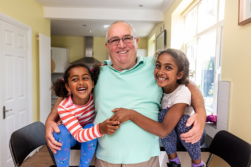 Waist-up shot of a senior male adult holding his granddaughters in each arm. He is looking into the camera while his granddaughters are both smiling with him. They are standing in the family kitchen.