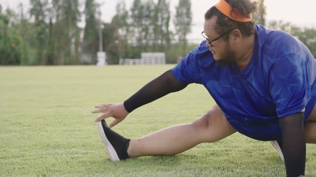 Overweight Asian man with very big belly sitting and stretching both legs, and doing a warmup exercise on a grass field at a stadium before running exercise after work.