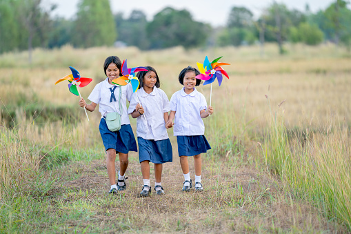Group of little Asian girls hold small windmill and look fun to run together in walkway of rice field.