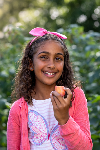 Waist-up shot of a young girl looking into the camera standing outdoors in a garden. She is about to place a freshly picked apricot in her mouth.