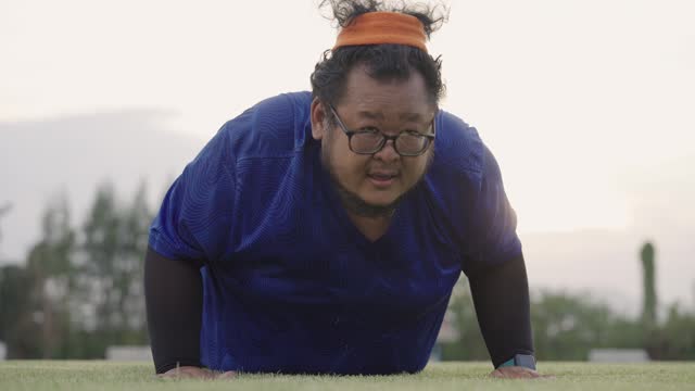 An obese Asian man putting a lot of effort to do push-ups on the grass field at a stadium in the evening as his routine for weight loss purpose.