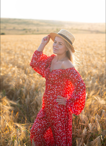 caucasian blonde woman in a red dress and hat on the golden wheat field in sunset light