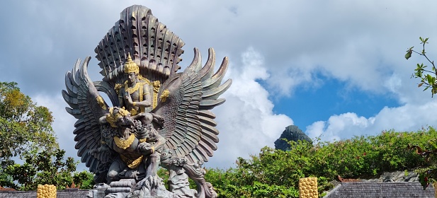 Ungasan, Bali - July 16, 2023: Wide picture of Hinduism God Vishnu and Garuda statue in Ungasan garden area with greenery plants, blue sky and white cloud as the background