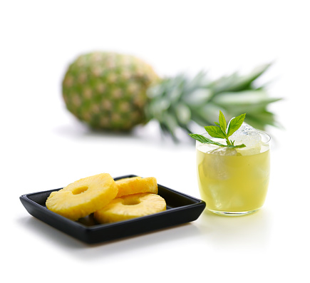 Sliced pineapple kept on a wooden table besides a glass filled with pineapple juice and a glass bowl with pineapple slices