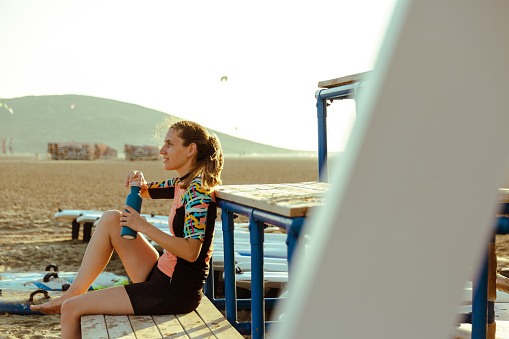 A young Caucasian female surfer is taking a break and opening her bottle of water.