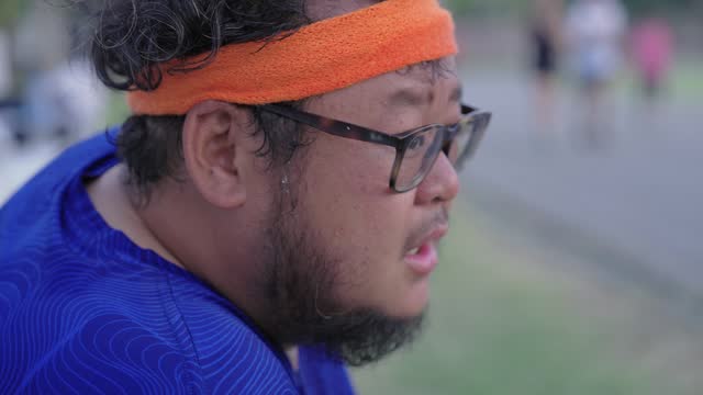 Side view of an exhausted chubby Asian man sitting on a bench while taking some rest after exercising. He's taking off his orange headband and rubbing sweaty eyes and face and looking at other people who are exercising at a stadium.