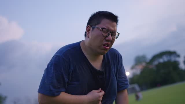 Close-up of a chubby Asian man with blue t-shirt drenched in sweat and panting hard with extreme exhaustion, standing on the soccer field while drinking some bottled water and pouring it onto his head to cool off the heat.