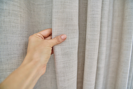 Light beige sand linen natural curtains on the window, close-up of a hand touching the curtains. Textile window decoration, fashion, style, design concept