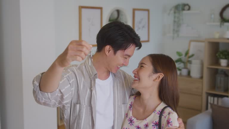 Happy young Asian couple smiling and showing keys to their new house they bought together preparing to get married and start a family.