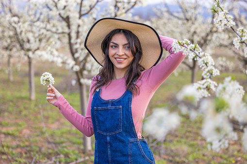 Pretty girl poses with hat on her head posing in blooming plum orchard in spring and holding flowers in her hand.