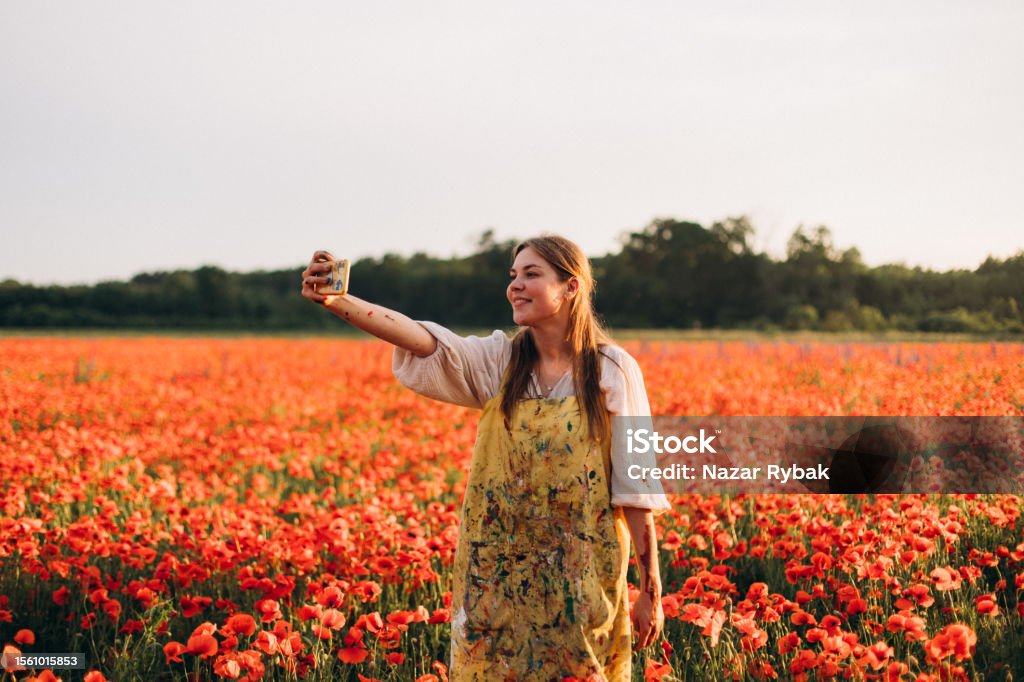 The cheerful woman doing a selfie on the scenic poppies field at the sunset In this cheerful and captivating scene, a woman finds joy and excitement amidst a scenic field of red poppies during the enchanting sunset. With a beaming smile, she holds up her smartphone and captures a selfie to commemorate the beautiful moment. Adults Only Stock Photo