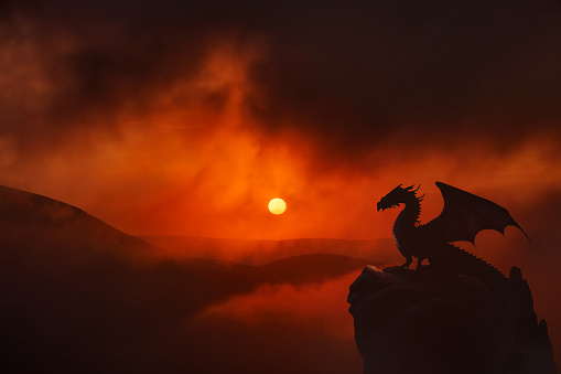 Mountain landscape with dragon winged silhouette sitting on the rock. Dramatic sunset in clouds background